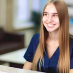 mujer de 22 años de edad, cabello largo y liso color rubio, enfermera de traje color azul oscuro, sonriendo, sentada en la recepción con fondo de hosp